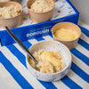 Borough 2 gluten free plant-based dessert cups shown with their blue branded box, arranged on a blue and white striped placemat. One cup is in the foreground topped with custard, and another plain cup sits beside it.